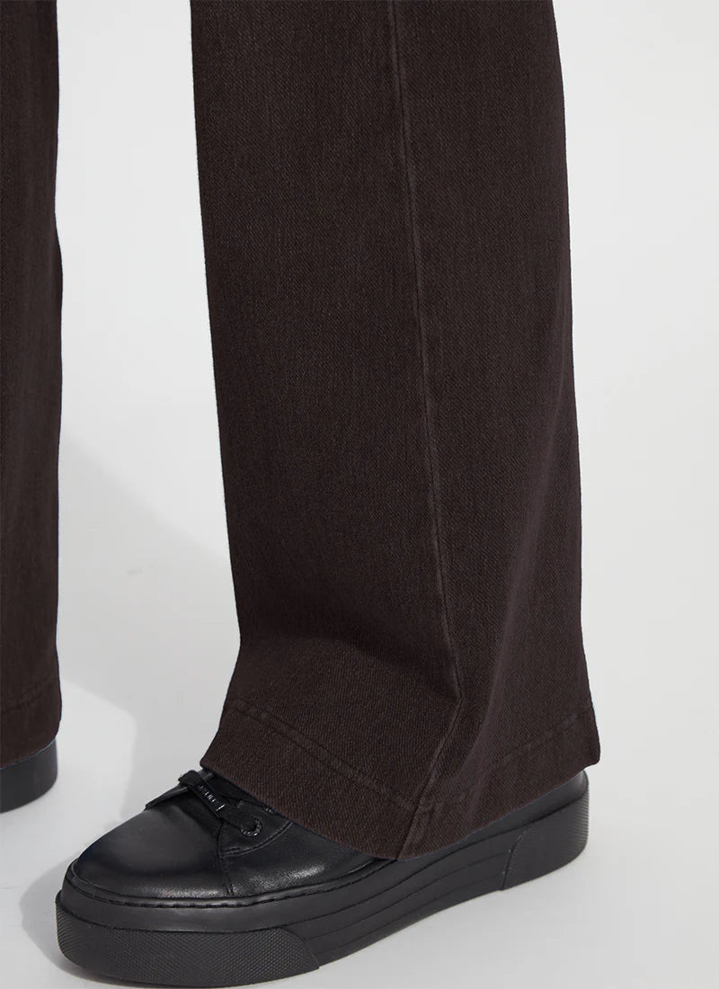 Close-up of a person's leg wearing dark brown wide-leg trousers and black platform sneakers against a light gray background.