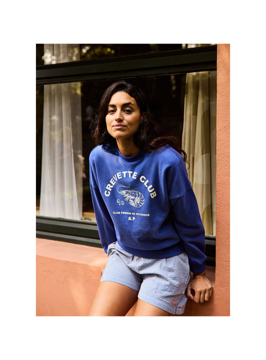 Person wearing a blue 'Crevette Club' sweatshirt standing in front of a window.