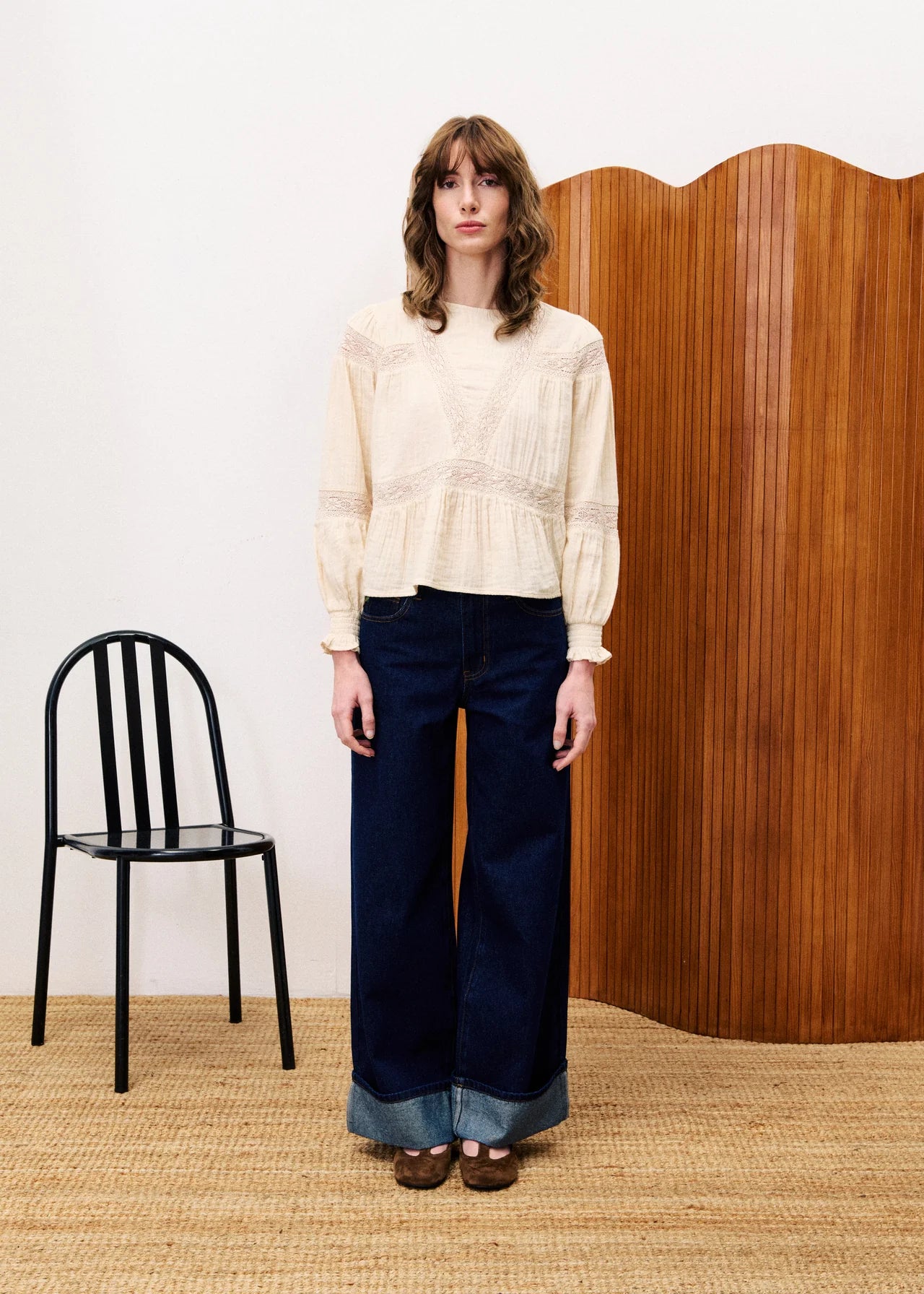 Woman wearing a cream blouse and navy pants standing in front of a wooden screen with a black chair on a beige carpet.