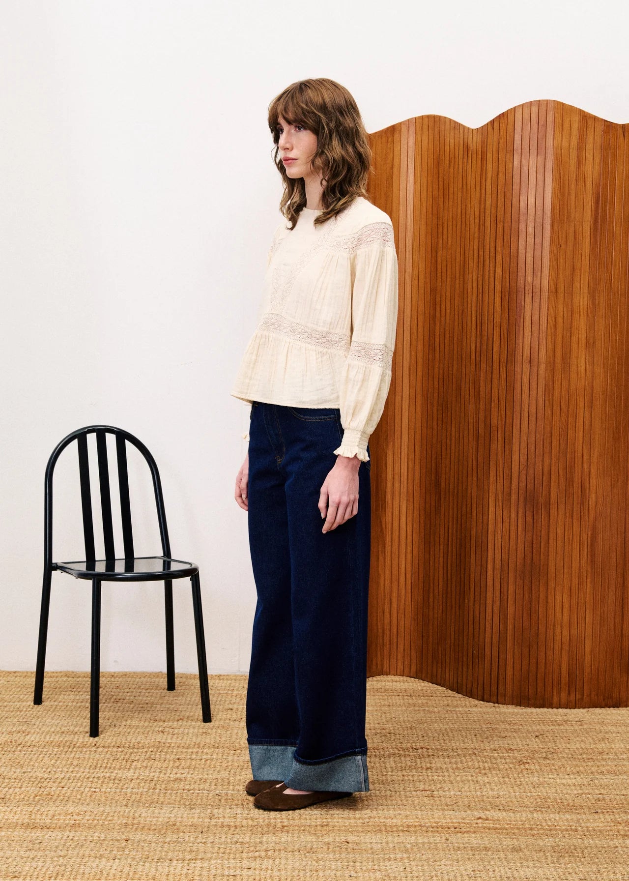 Woman in a white blouse and dark pants standing next to a black chair and wooden screen.
