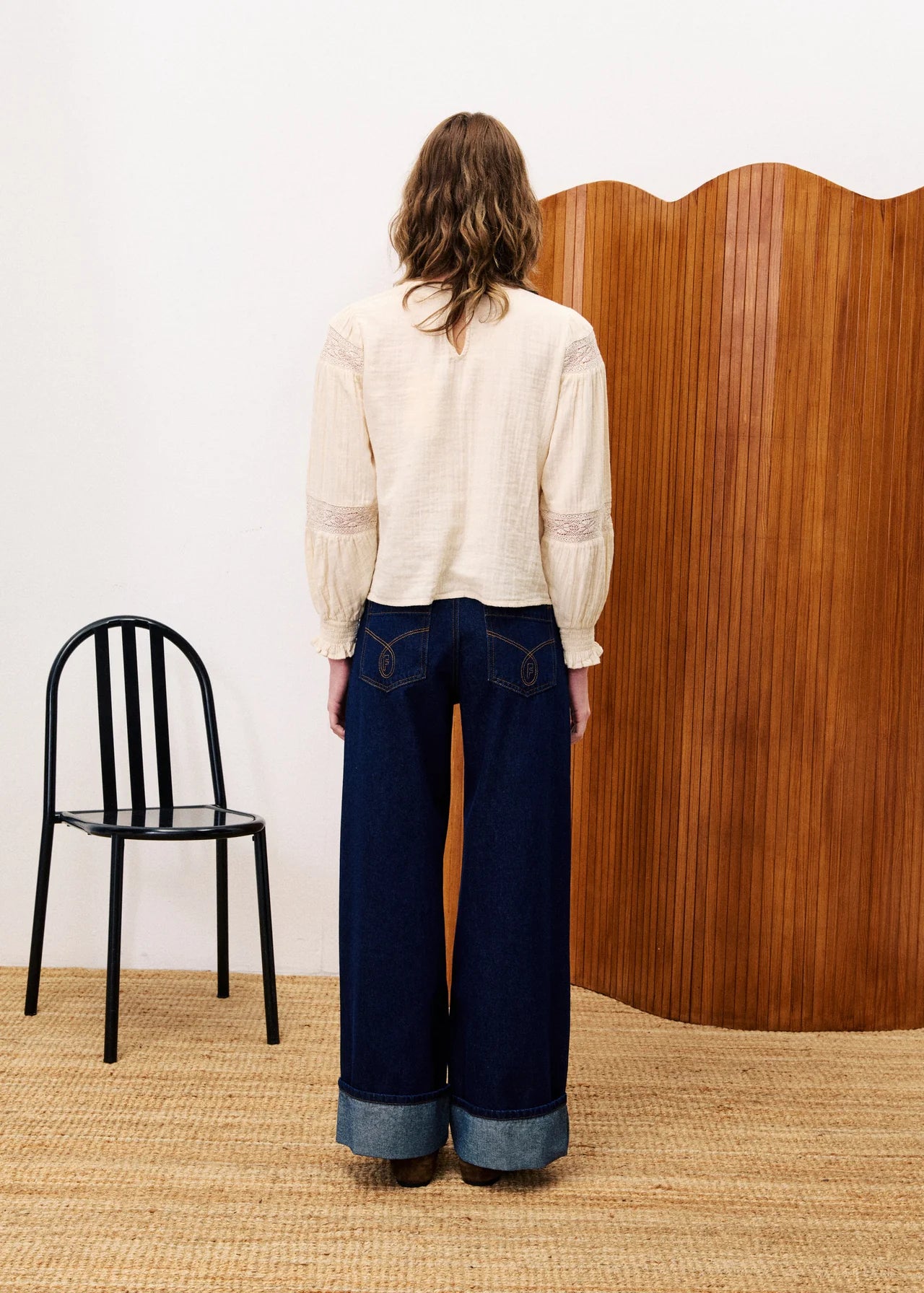 Person wearing a white blouse and dark blue jeans standing in front of a wooden screen with a black chair on a beige carpet.