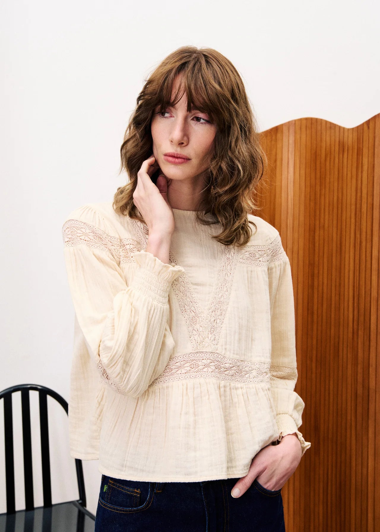 Woman wearing a cream lace blouse against a white wall with a wooden panel.