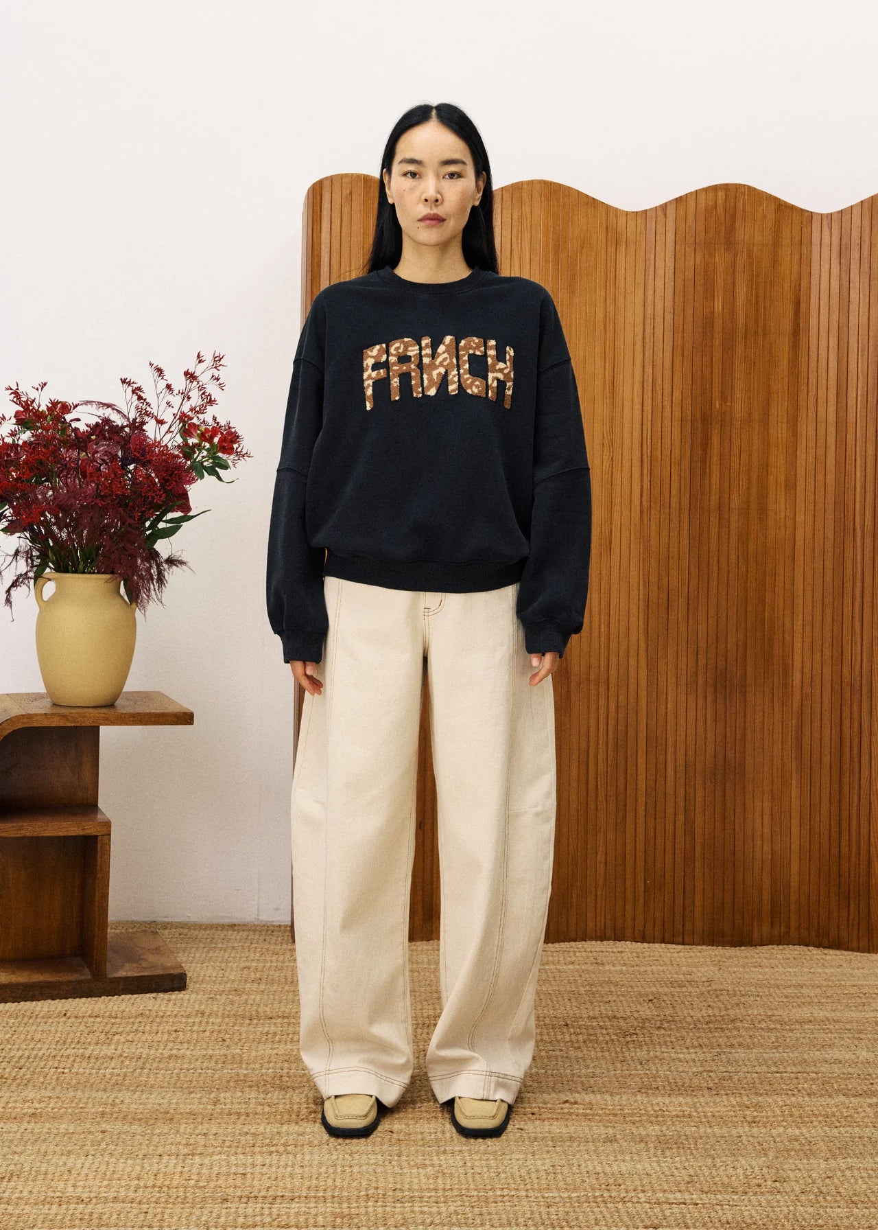 Person wearing a black sweater with 'FRNCH' text, standing in a room with wooden paneling and a vase of flowers.