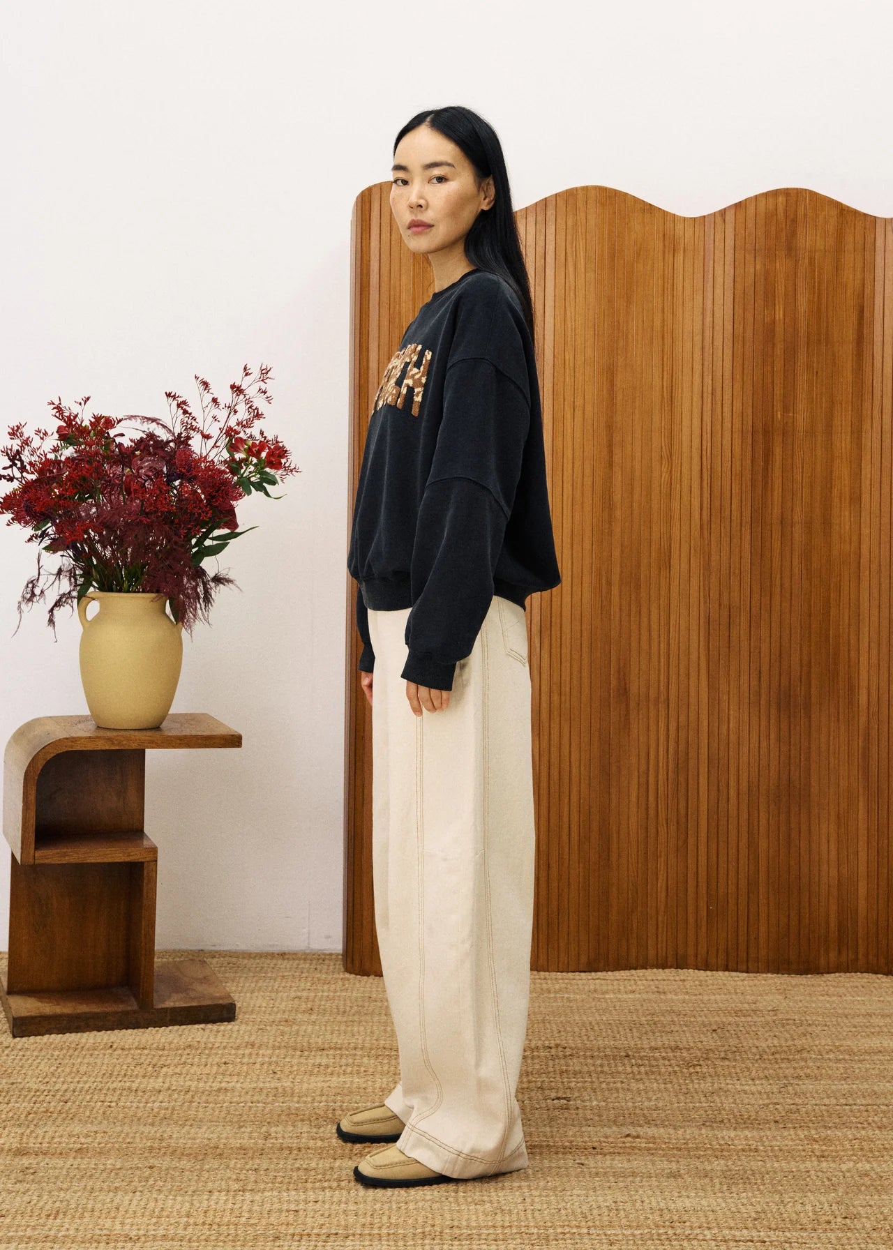 Woman standing in a room with wooden furniture and a vase of flowers.
