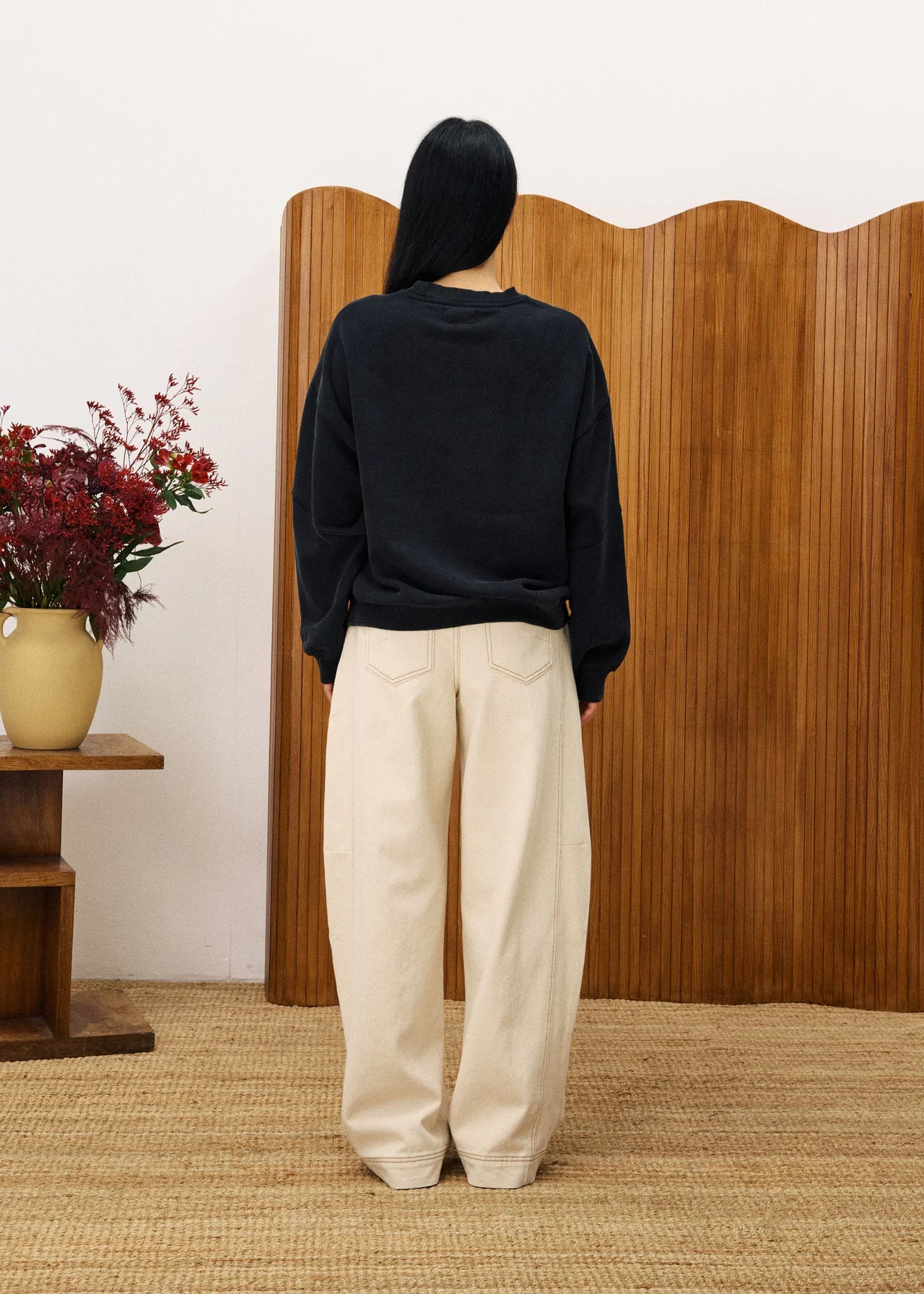 Person wearing a black sweater and beige pants standing in front of a wooden screen with a vase of flowers on a table.