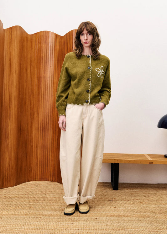 Person wearing a green sweater with floral design and beige pants standing in a room with wooden paneling.