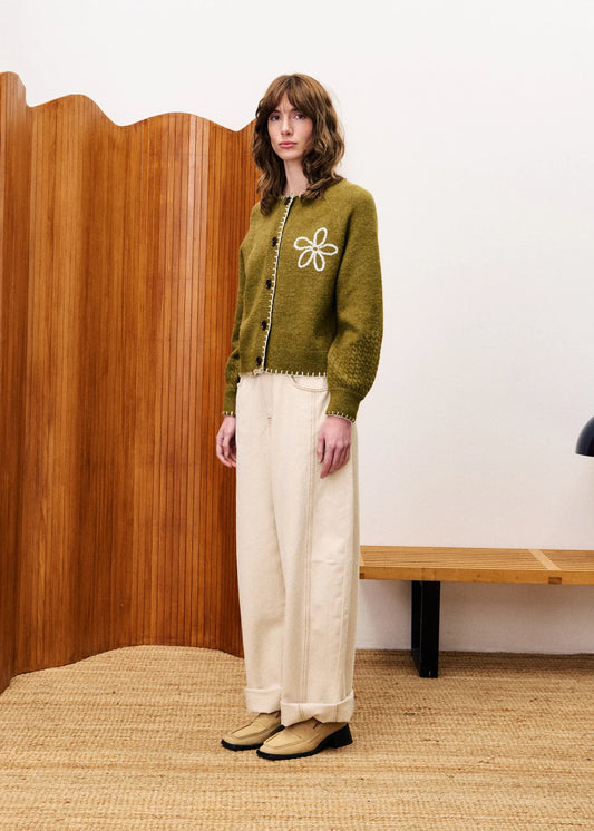 Person wearing a green sweater with a white flower design, standing in a room with wooden paneling and a white wall.