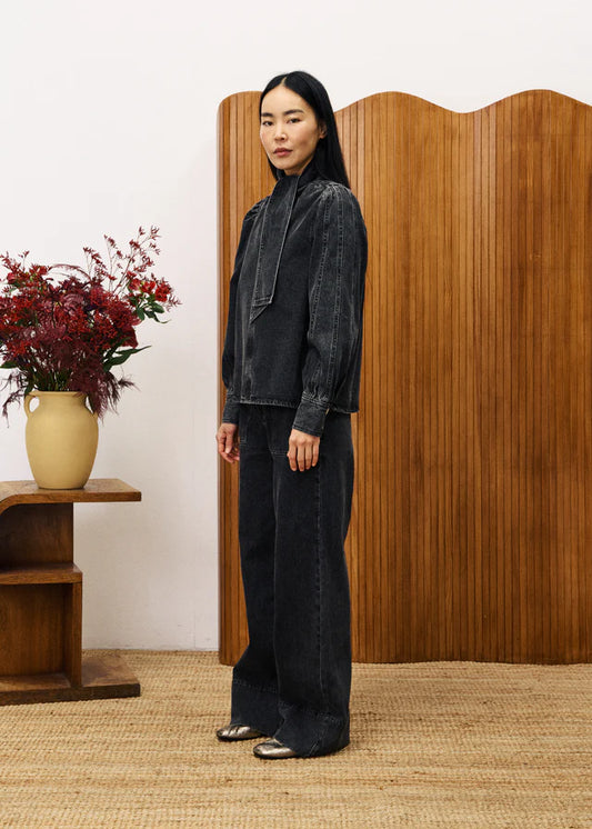 Person wearing a dark denim outfit standing in front of a wooden screen with a vase of flowers on a table.