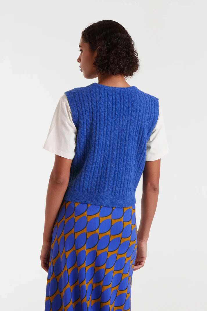 Person wearing a blue knitted vest over a white shirt with a patterned skirt on a white background