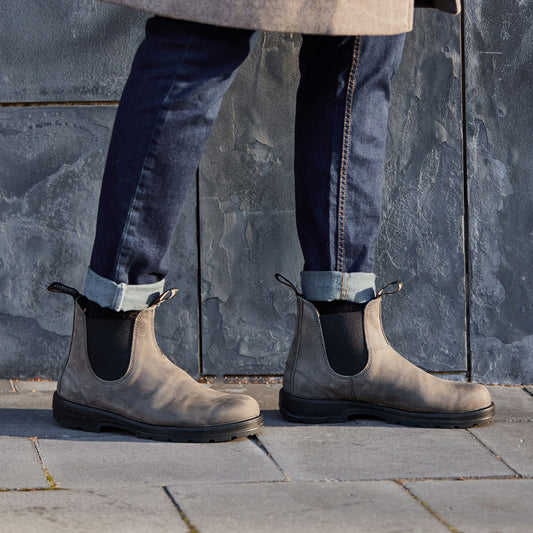 Person wearing gray Chelsea boots and dark jeans against a stone wall.