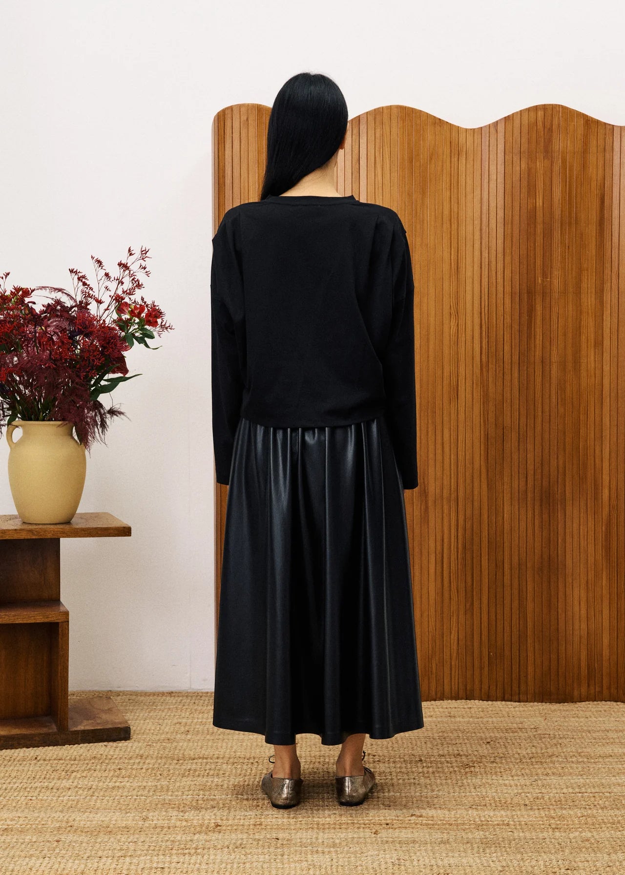 Person wearing a black outfit standing in front of a wooden screen with a vase of flowers on a small table.