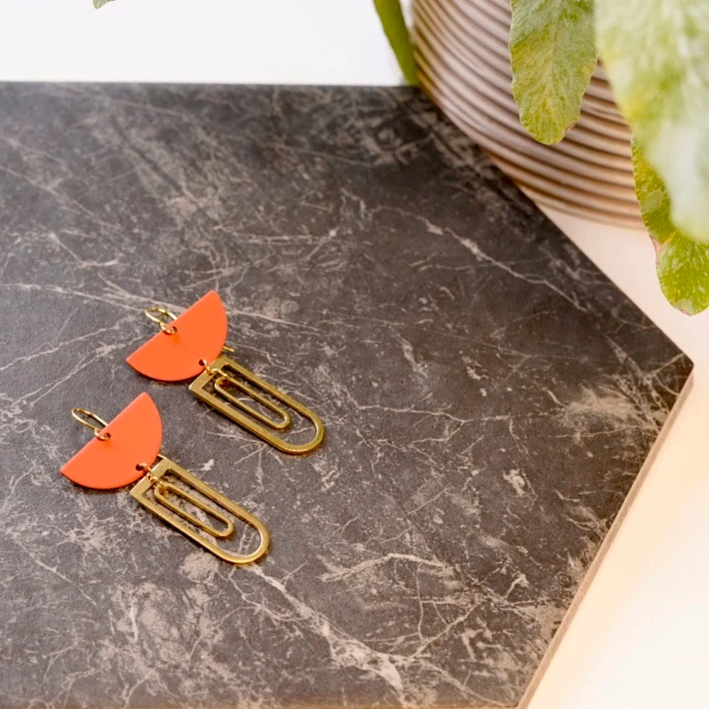 Pair of earrings with orange half-circle and gold bar design on a marble surface.