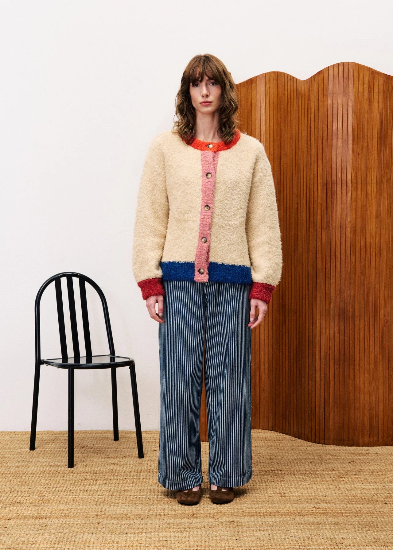 Person wearing a cream cardigan with colorful buttons, standing in a room with a black chair and wooden wall.