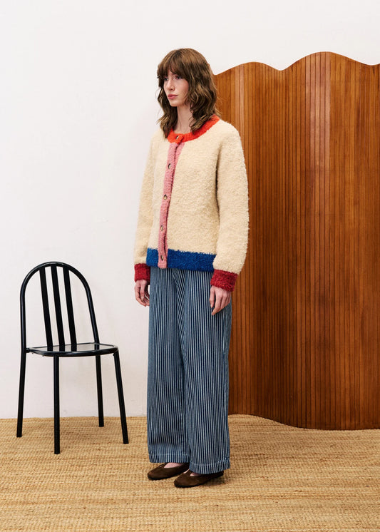 Woman wearing a colorful cardigan and striped pants standing in a room with a black chair and wooden wall.