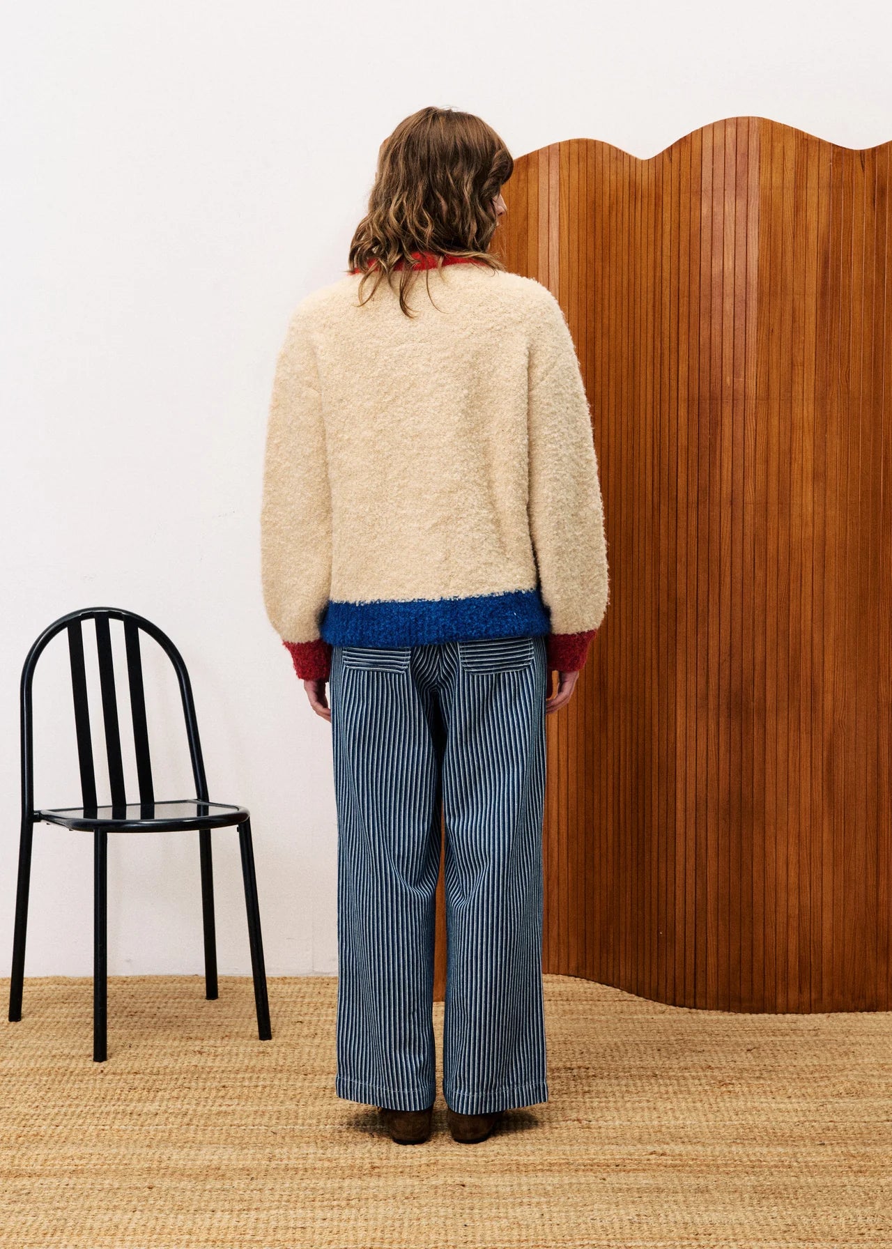 Person wearing a beige sweater and blue pants standing in front of a wooden screen with a black chair to the left.