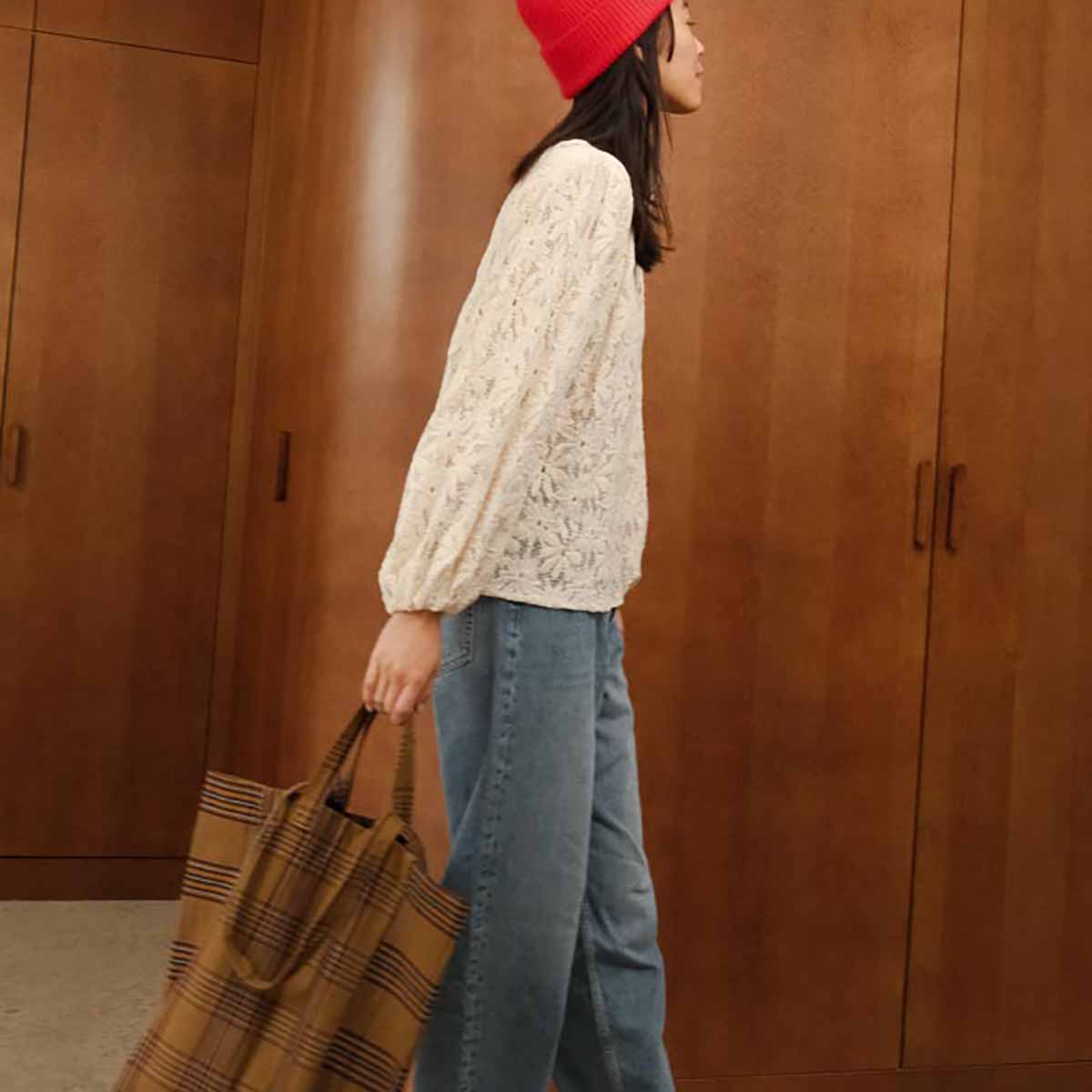 Person wearing a red beanie and white lace top, holding a plaid bag against a wooden background