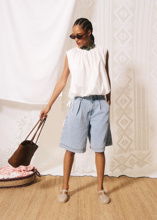 Woman in white sleeveless top and light blue shorts holding a woven bag against a textured white wall.