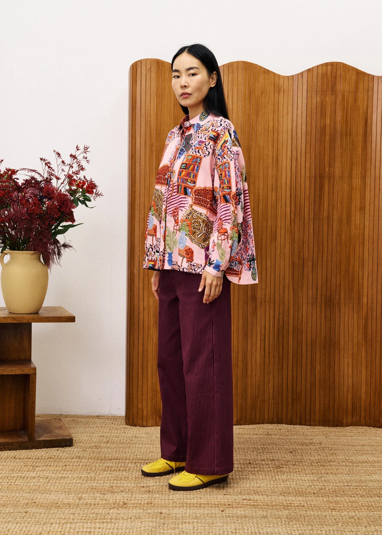 Woman wearing a colourful patterned blouse and burgundy pants standing in front of a wooden screen.