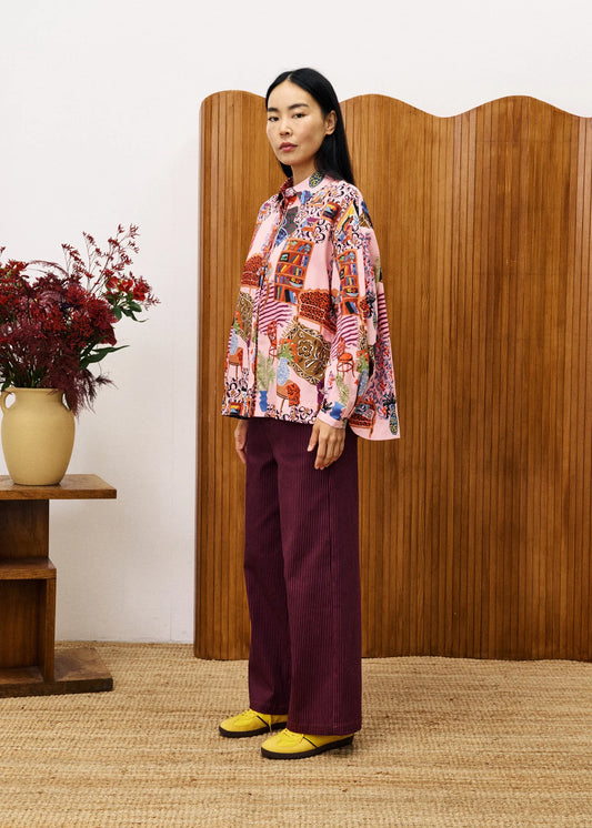 Woman wearing a colourful patterned blouse and burgundy pants standing in front of a wooden screen.