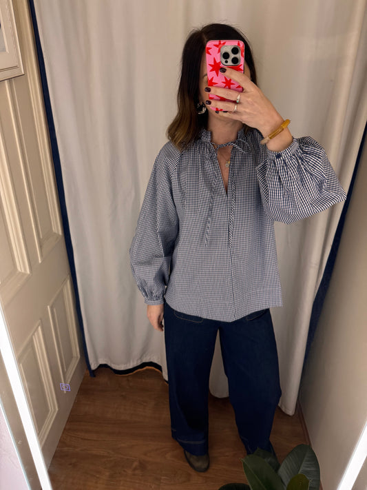 Person wearing a checkered blouse and dark pants in a room with curtains.