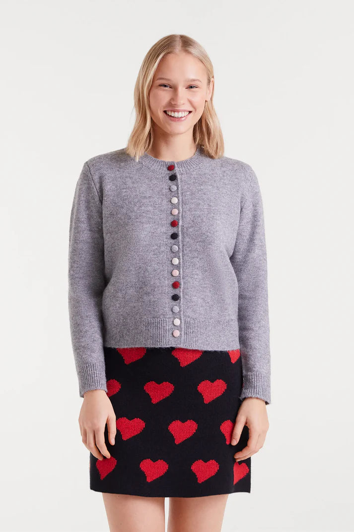 Woman wearing a gray cardigan with colorful buttons and a black skirt with red heart patterns on a white background