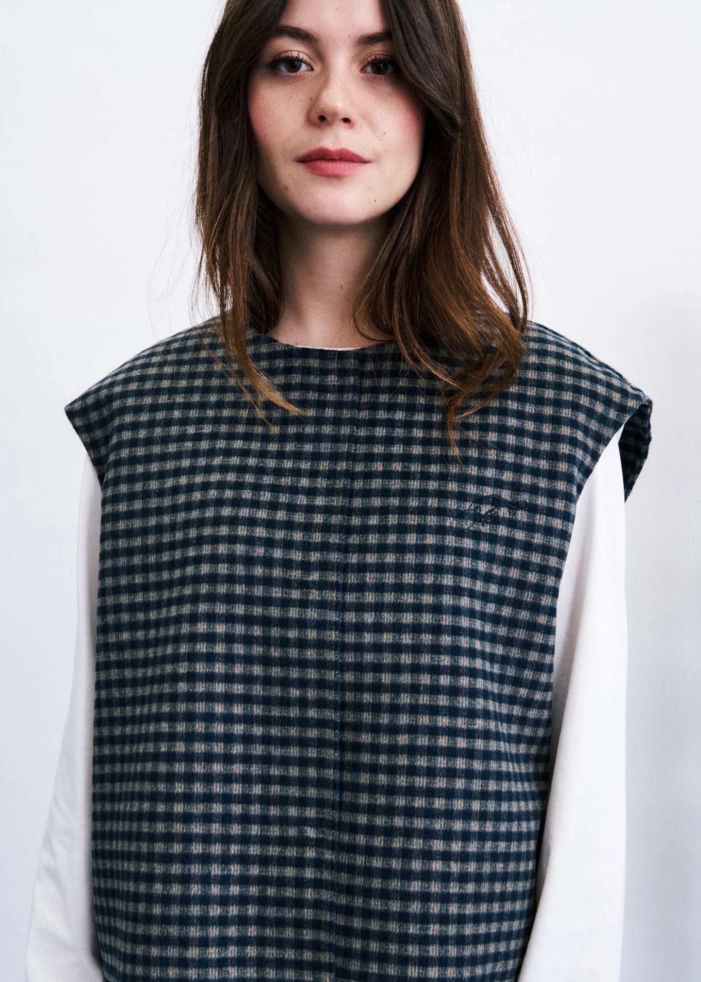 A woman wearing a sleeveless dark green and beige gingham patterned vest over a white long-sleeve shirt, photographed against a plain white background.