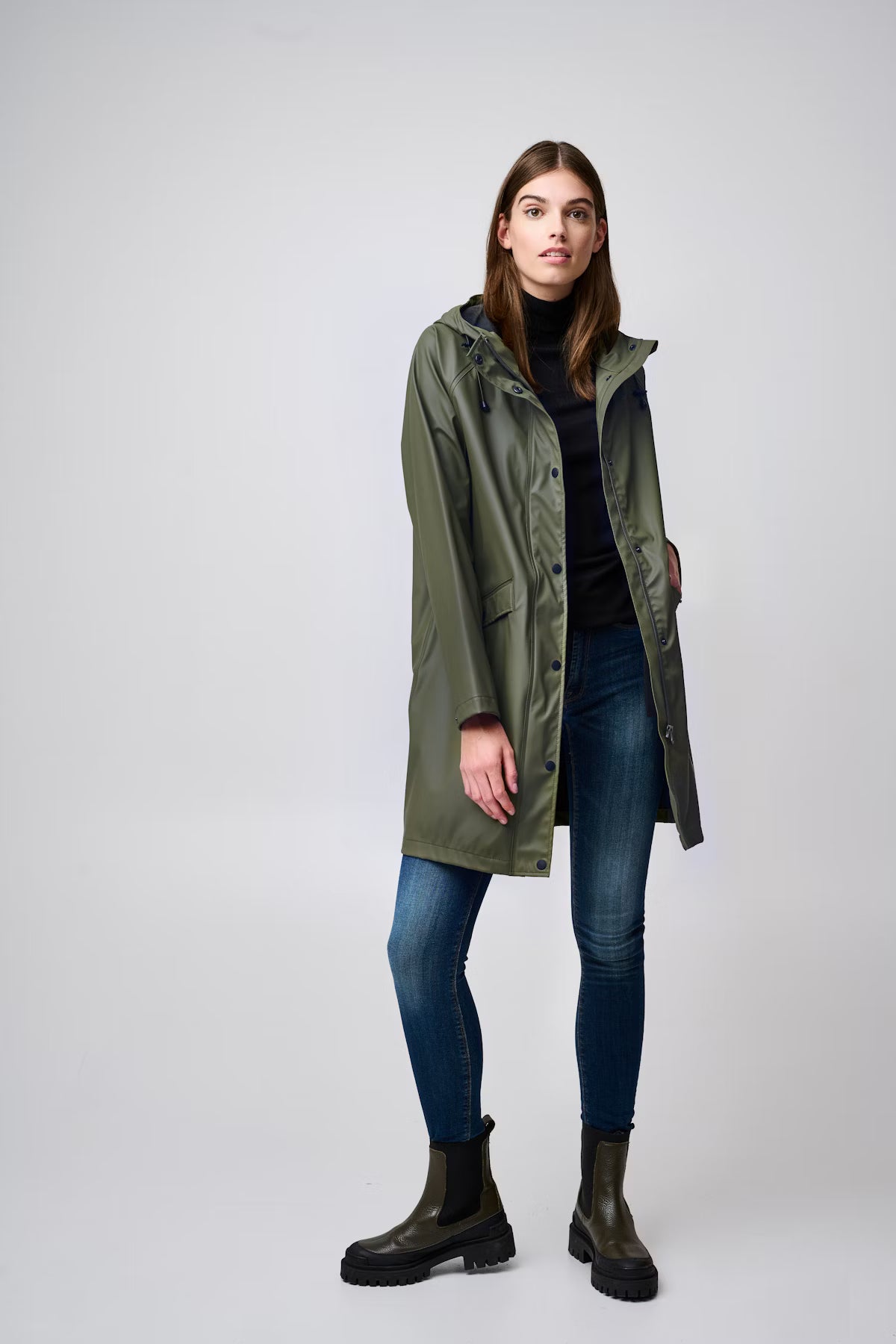 Woman wearing a green raincoat, black boots, and blue jeans on a white background