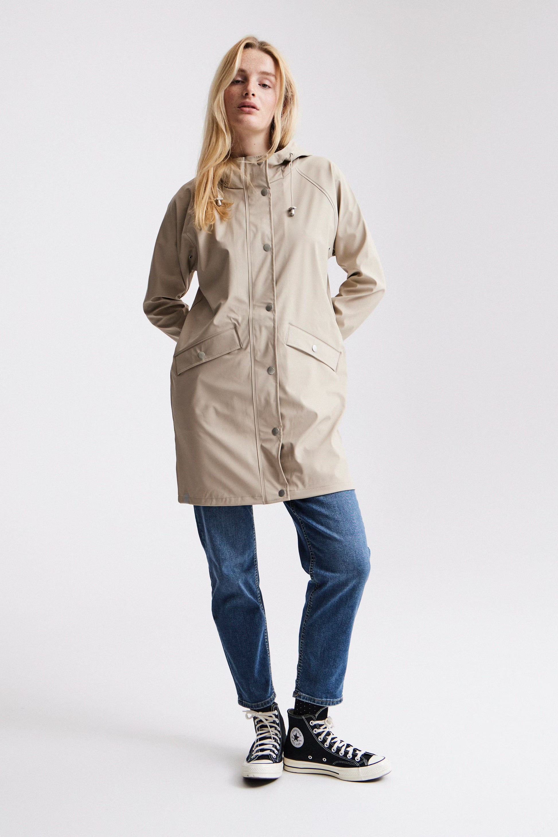 Woman wearing a beige raincoat, blue jeans, and black sneakers on a white background