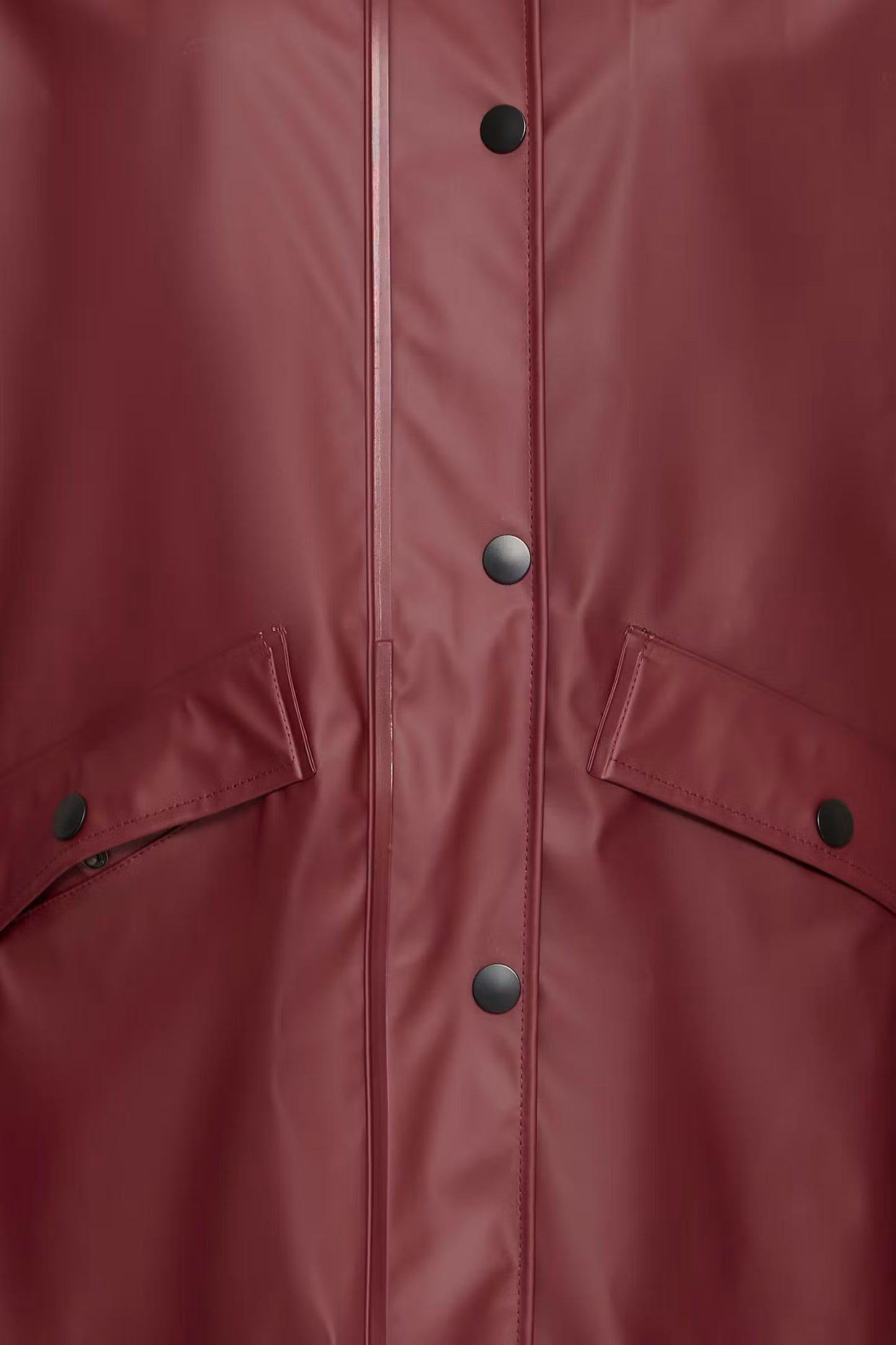 Close-up of a matte burgundy raincoat showing the front button closure, vertical seam, and flap pockets with snap buttons.