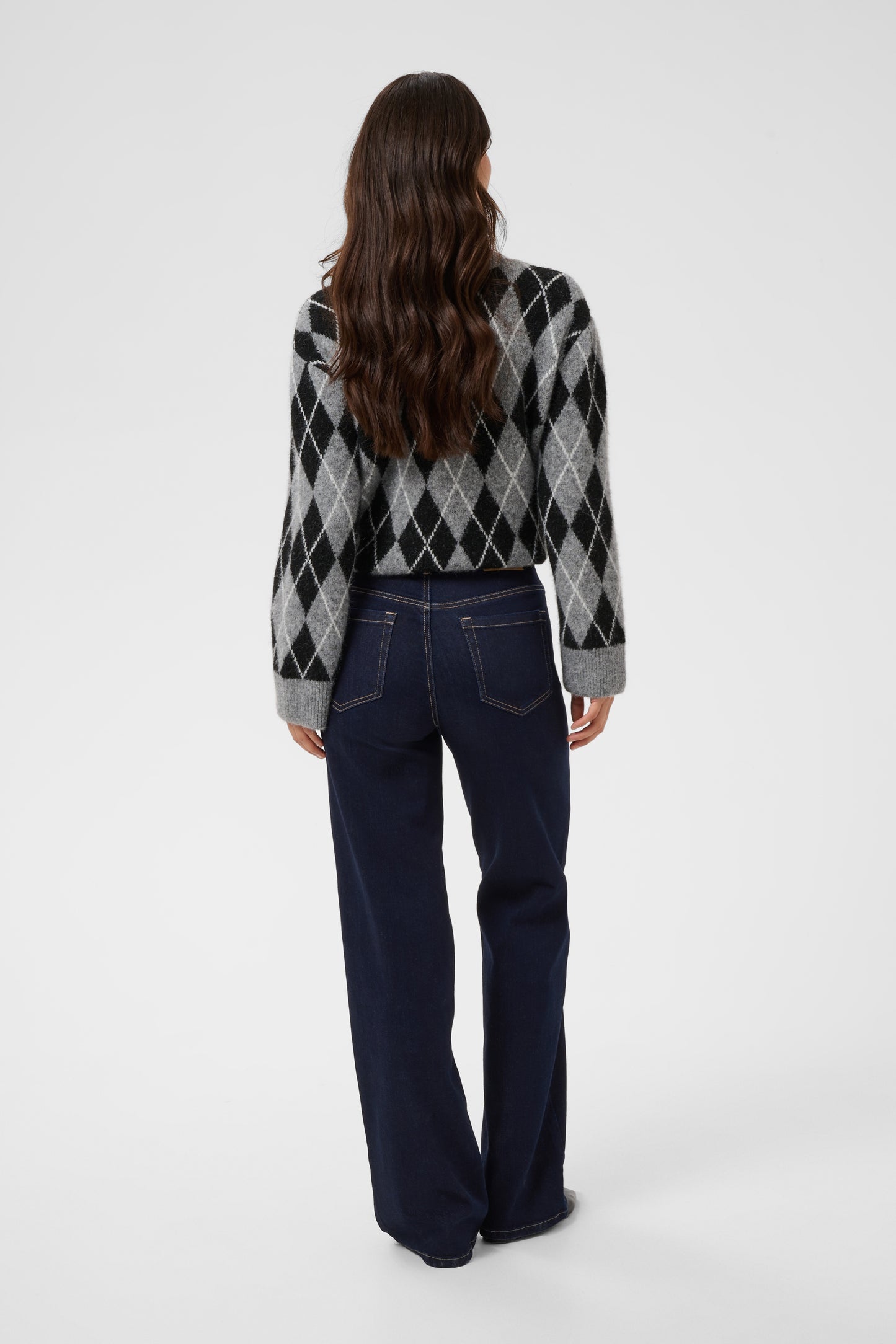 A woman viewed from the back wearing a gray, black, and white argyle sweater and dark blue wide-leg jeans against a light gray studio background.