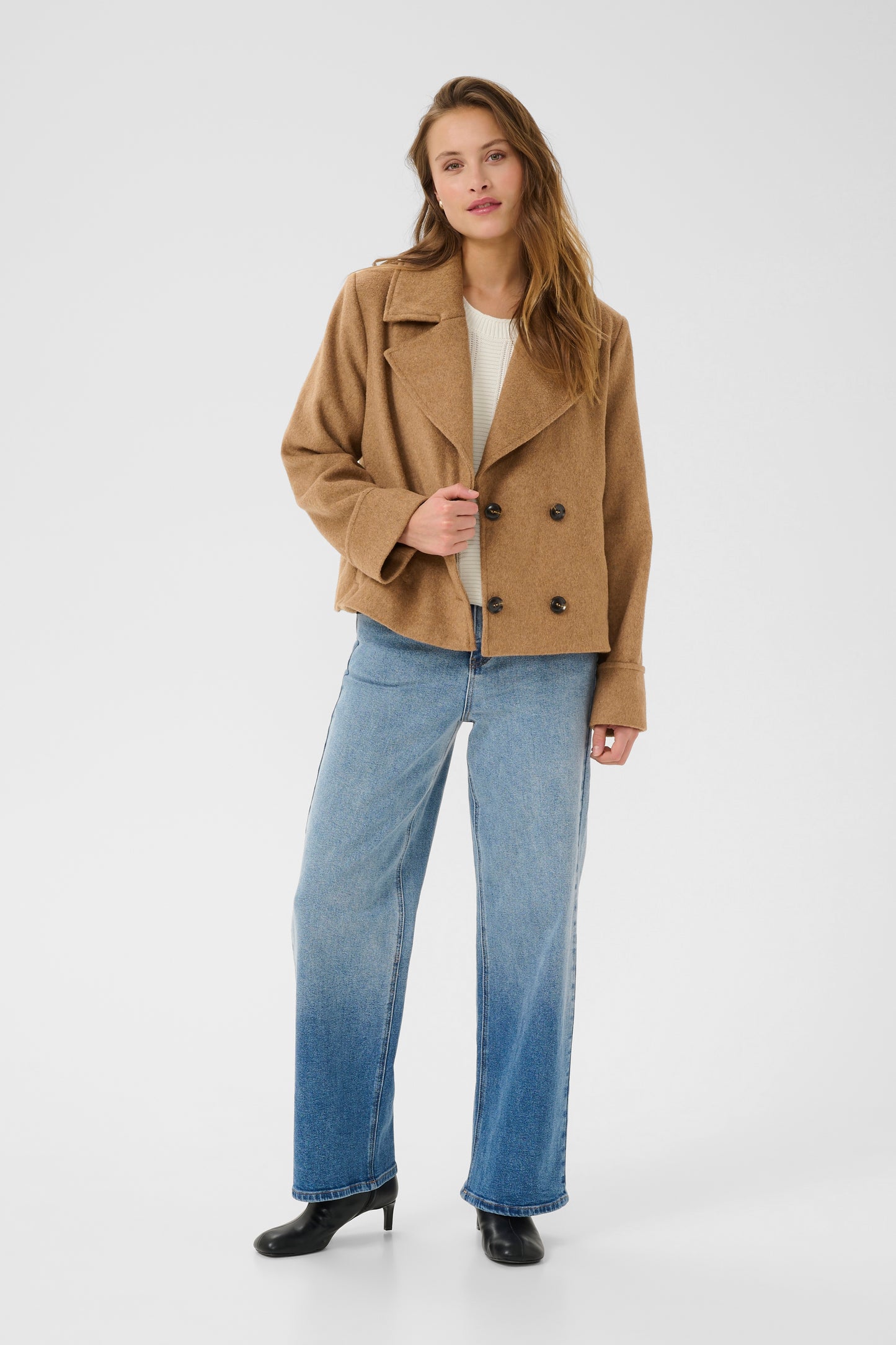 Woman wearing a tan coat, white shirt, and blue jeans on a white background