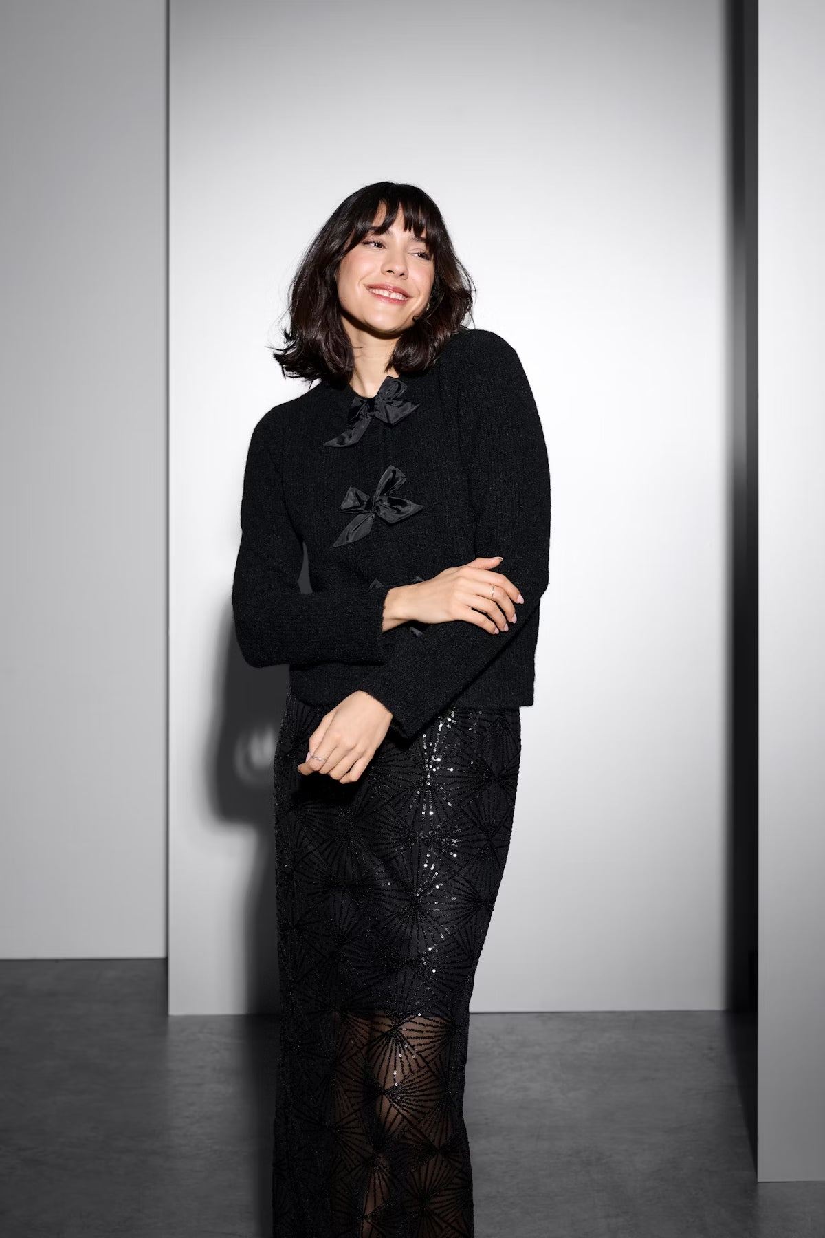 Woman wearing a black cardigan with a bow detail and a sequin skirt against a grey background