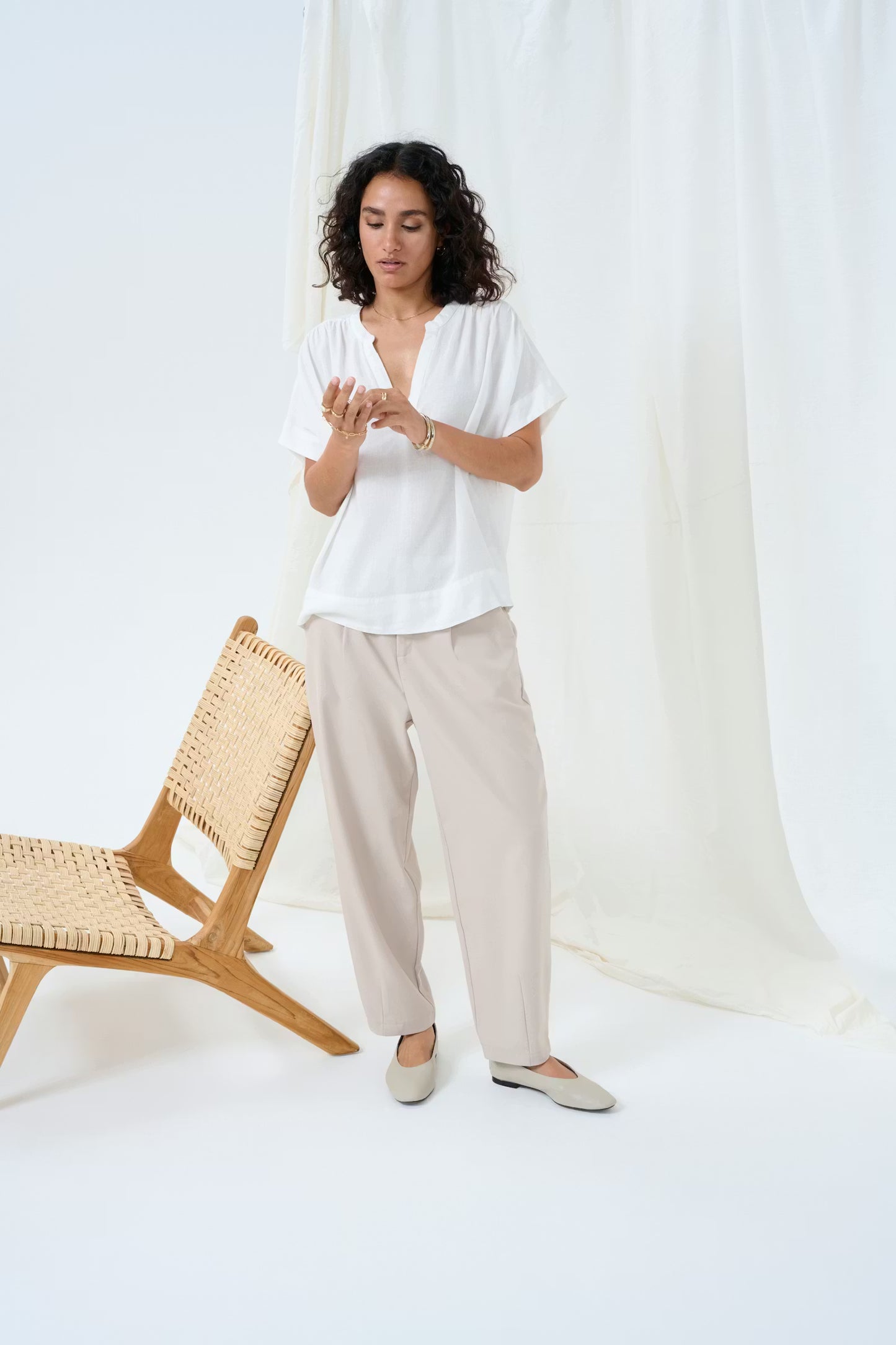 Woman in a white blouse and beige pants standing in a minimalistic room with a wooden chair.