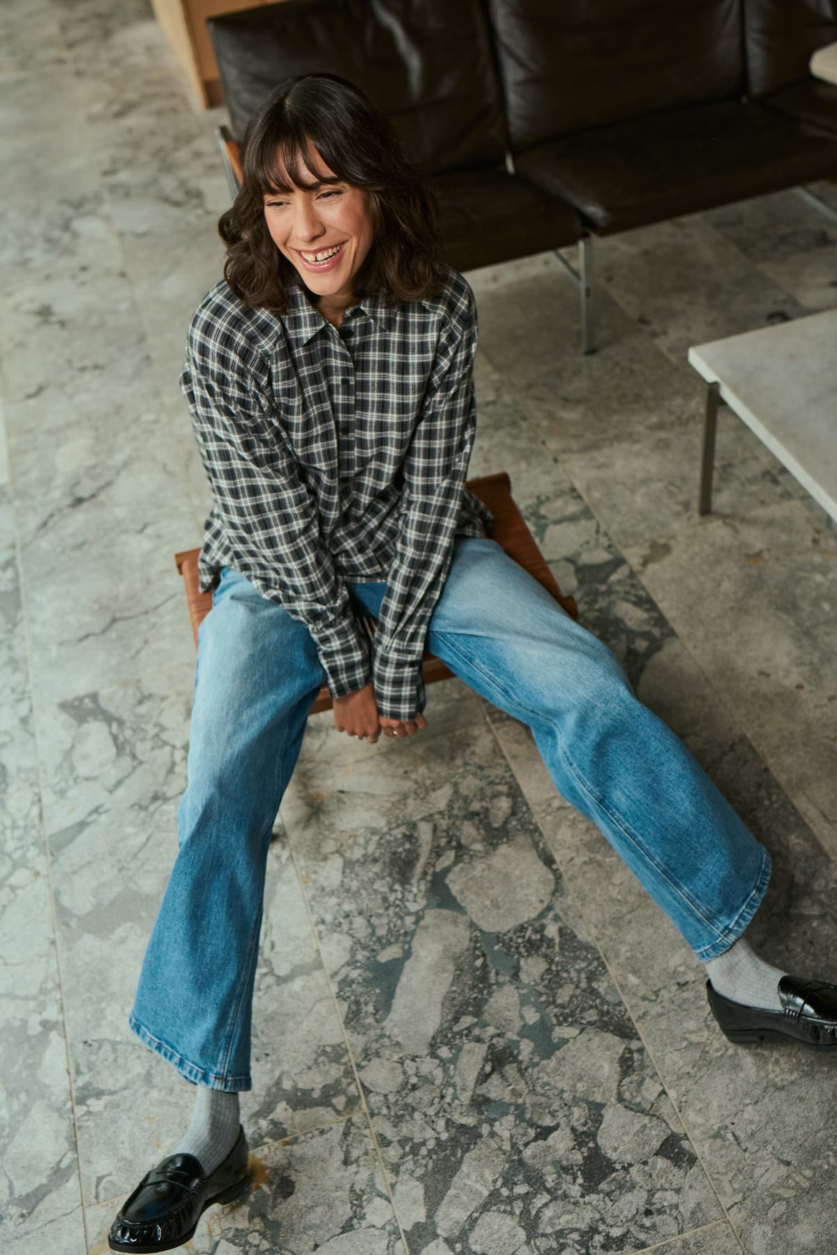 Person sitting on a chair wearing a checkered shirt and jeans in an indoor setting.