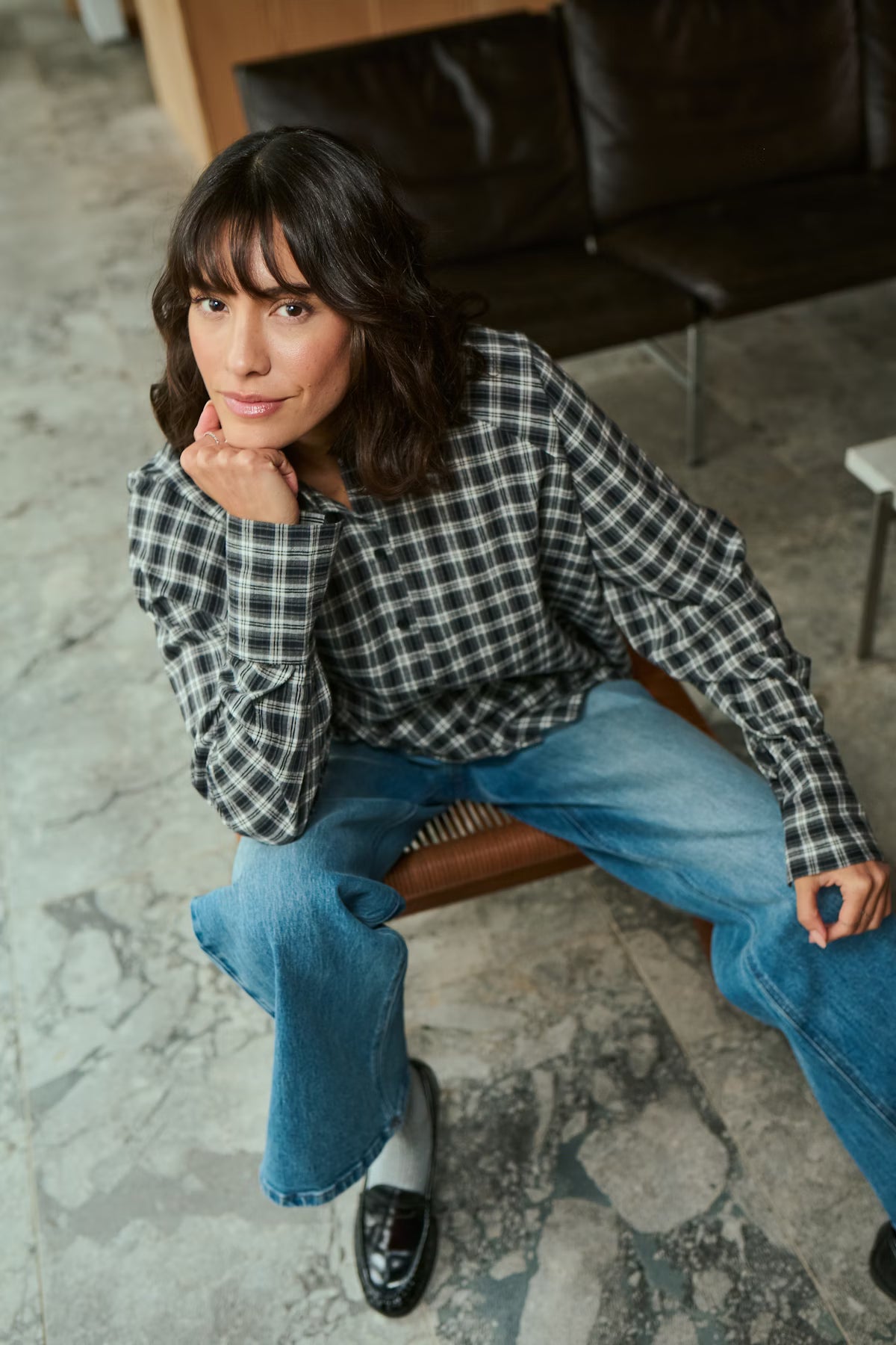 Woman sitting on a chair wearing a plaid shirt and jeans in an indoor setting.