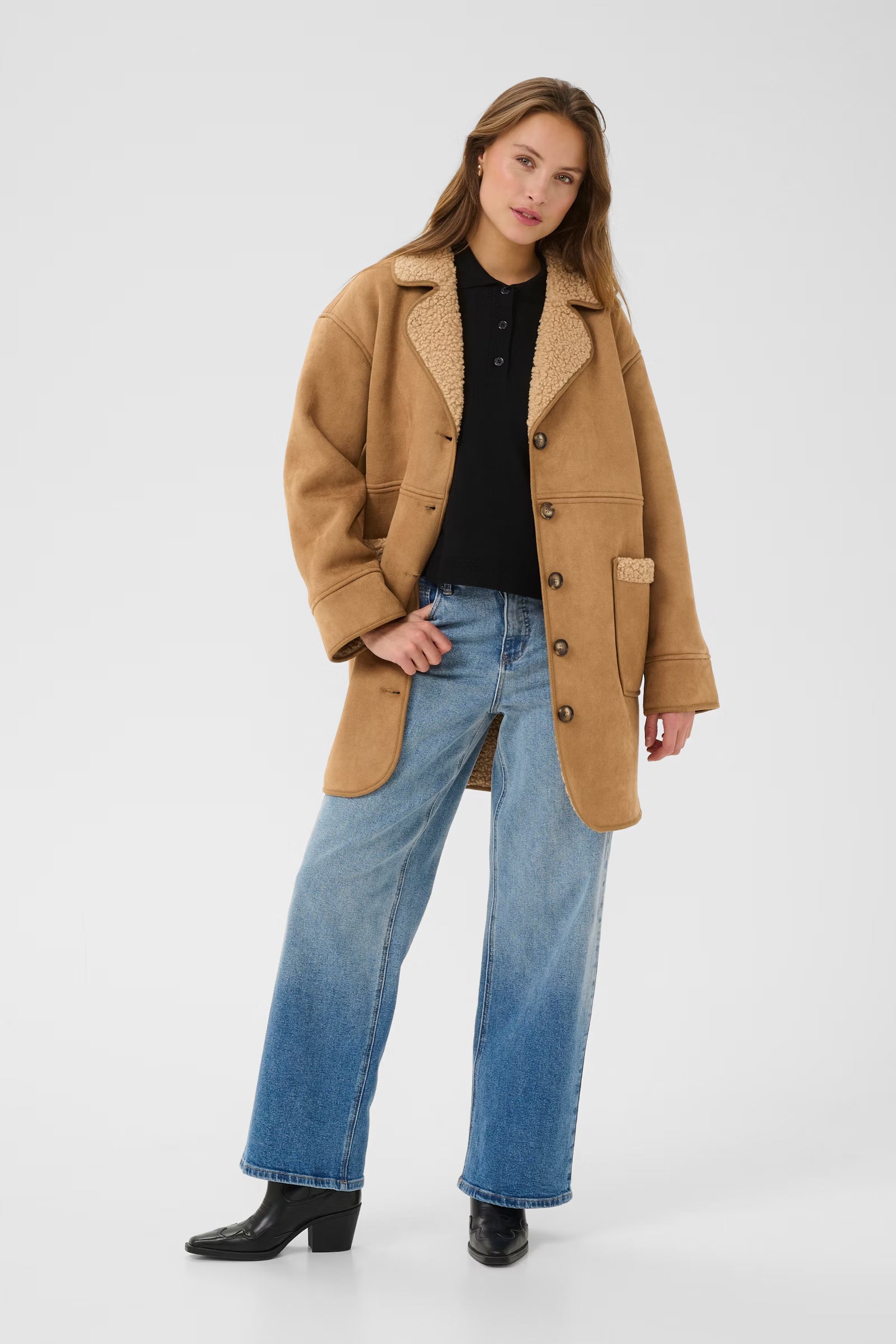Woman wearing a tan coat, black shirt, blue jeans, and black boots on a white background