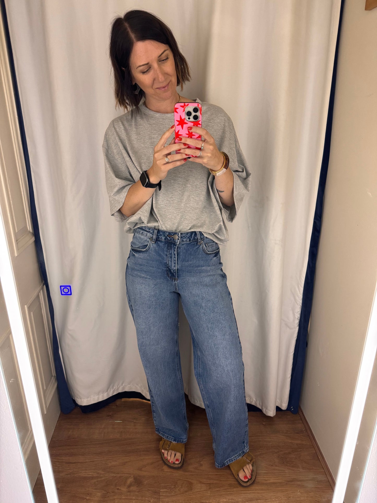 Woman taking a mirror selfie with a wooden floor and white curtain in the background.