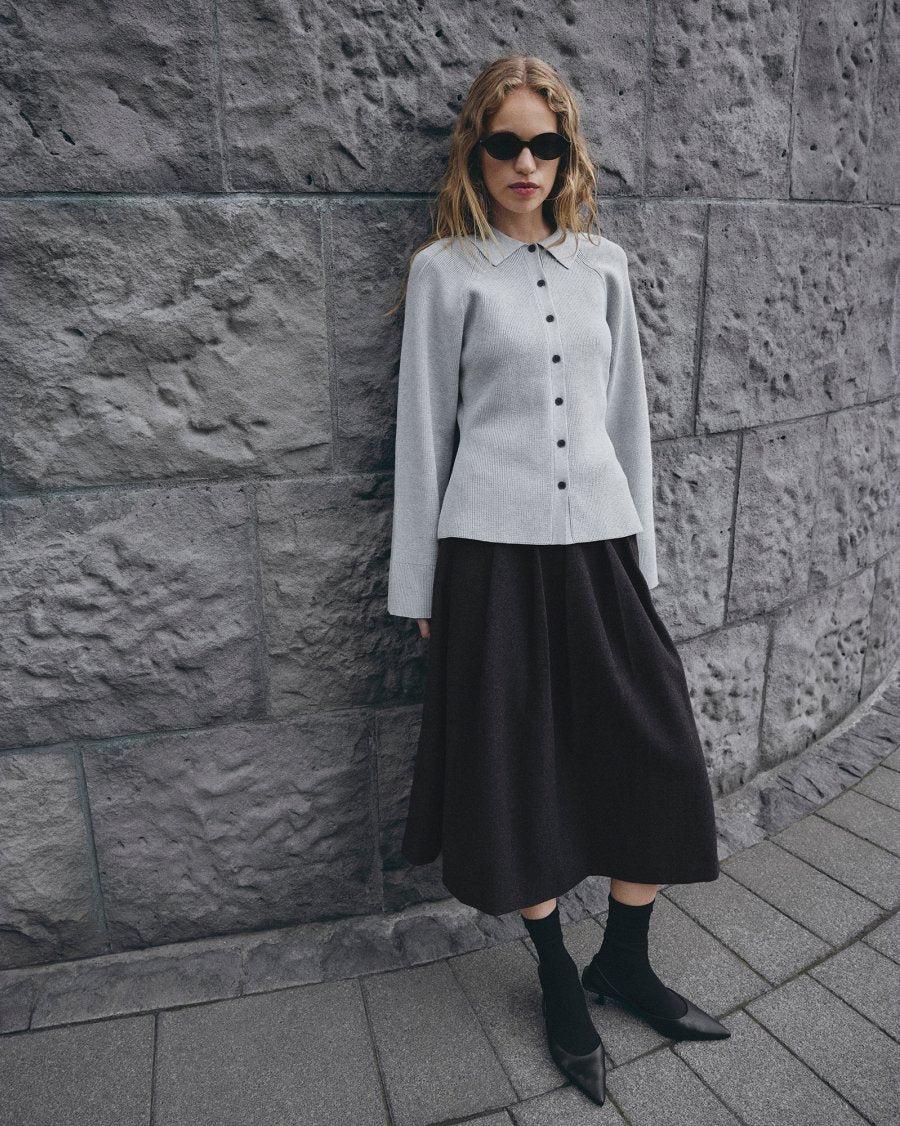 Woman wearing a light gray blouse and black skirt against a stone wall.
