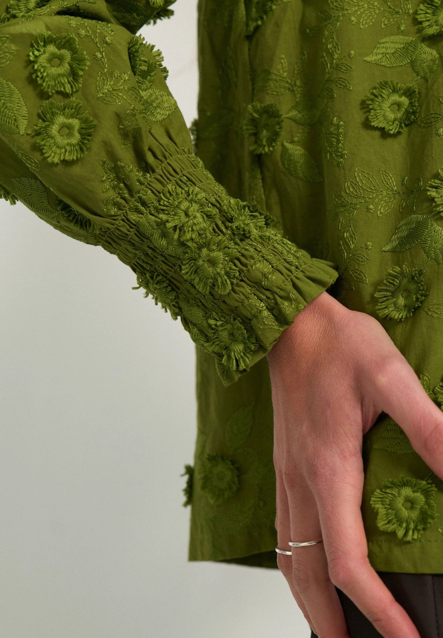 Close-up of a person wearing an olive green embroidered blouse with textured floral applique and smocked sleeves, with a hand resting near the waist.