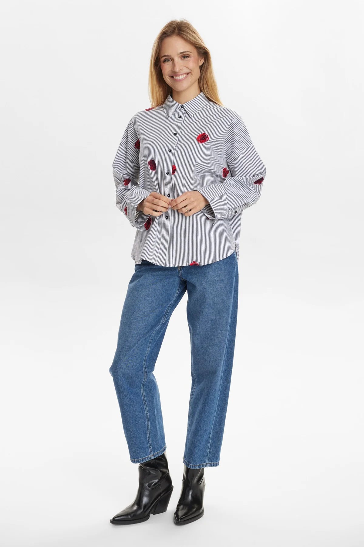 Woman wearing a stripe shirt with red floral patterns, blue jeans, and black boots on a white background