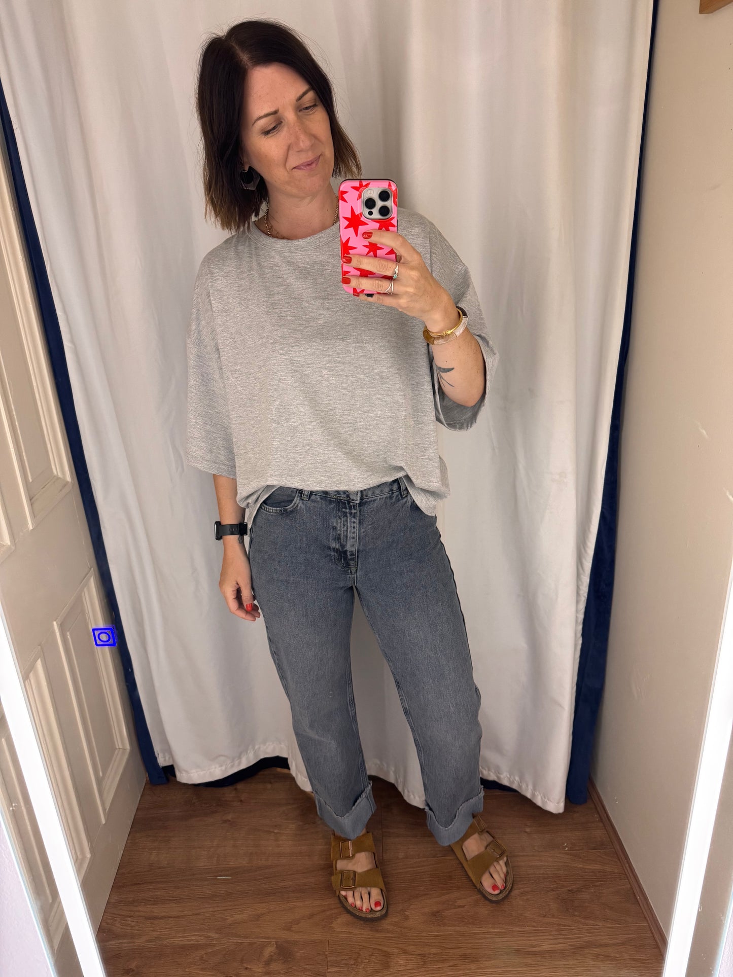 Woman taking a mirror selfie wearing a gray shirt and blue jeans indoors.