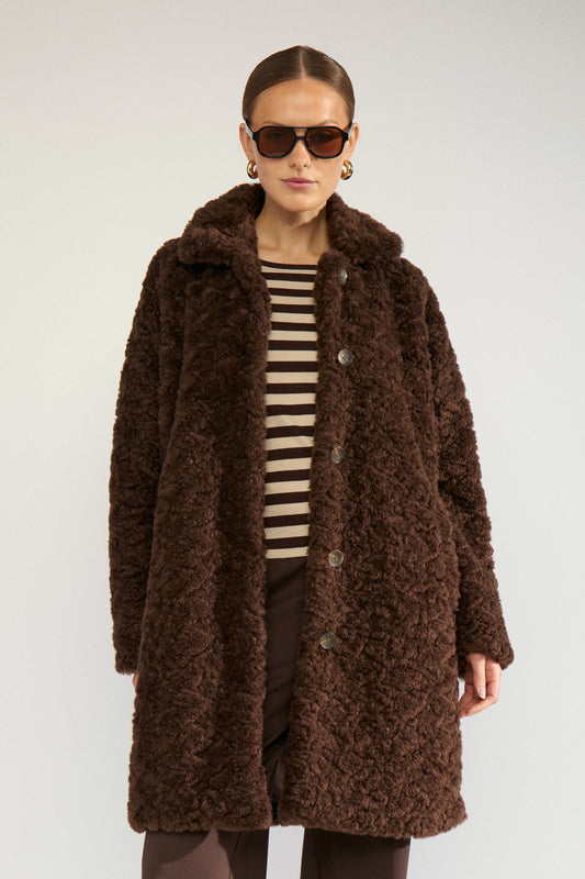 A woman modeling a brown textured teddy coat over a black and beige striped top, photographed against a plain light gray studio background.