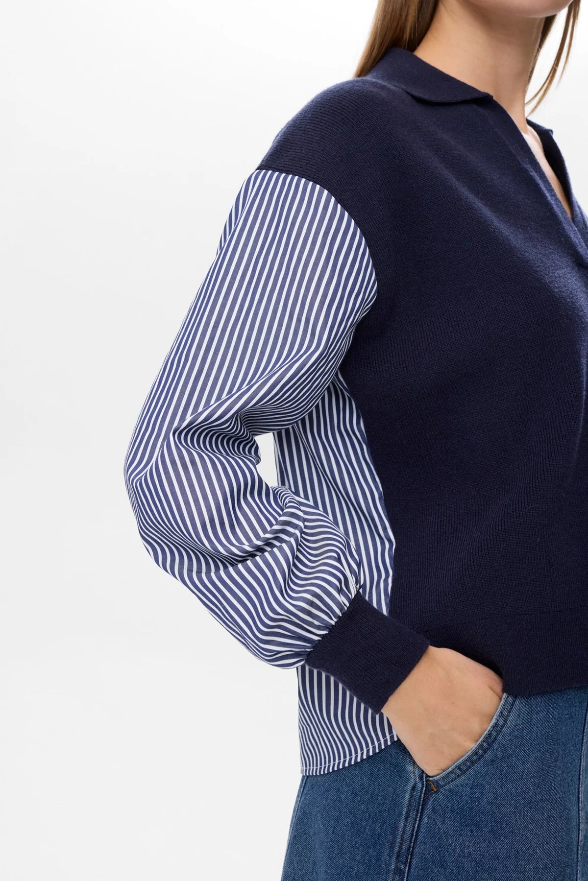 Person wearing a navy sweater over a striped shirt with blue jeans on a white background