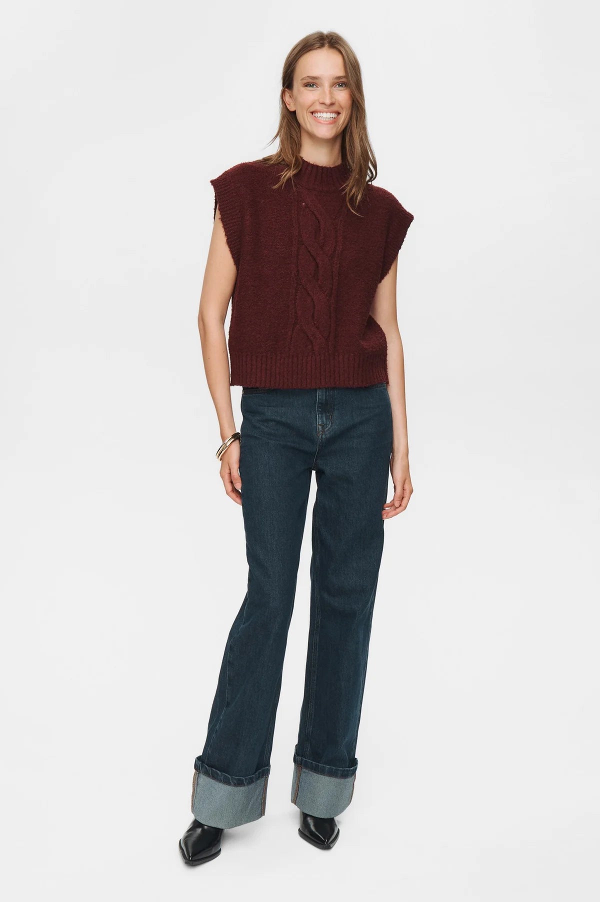 Woman wearing a burgundy sleeveless sweater and dark blue jeans on a white background