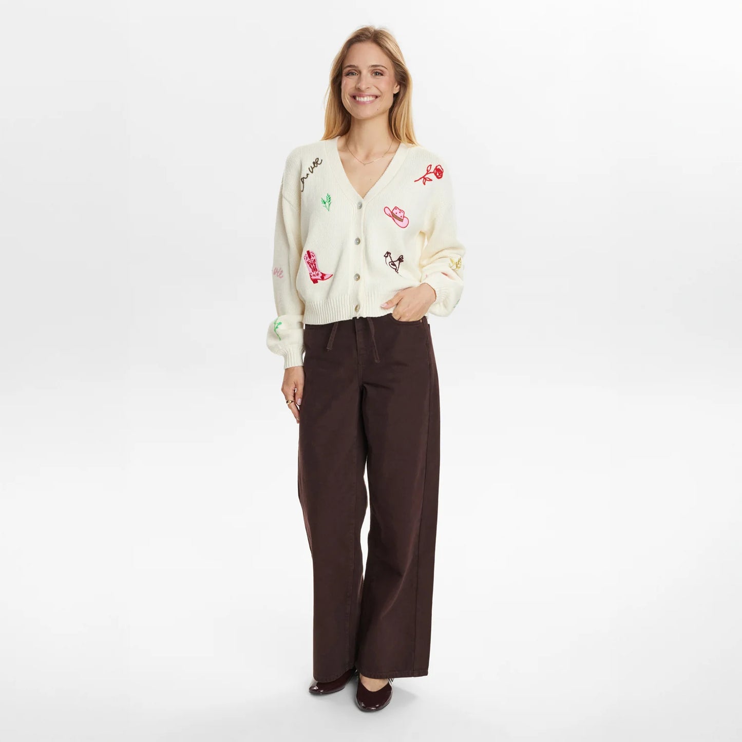 Woman wearing a cream cardigan with colorful embroidery and brown pants on a white background
