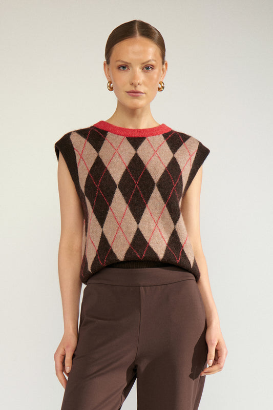 A woman modeling a sleeveless knit top with an argyle pattern in beige, brown, and red, paired with brown trousers, against a light gray studio background.
