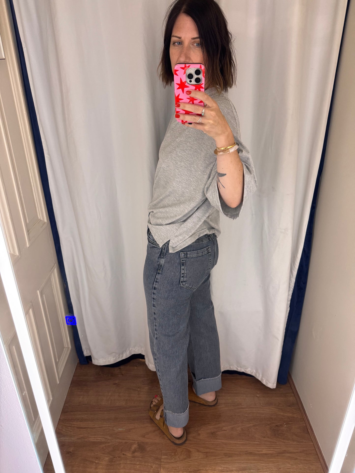 Person taking a mirror selfie wearing a grey t shirt and blue jeans in a room with white curtains.