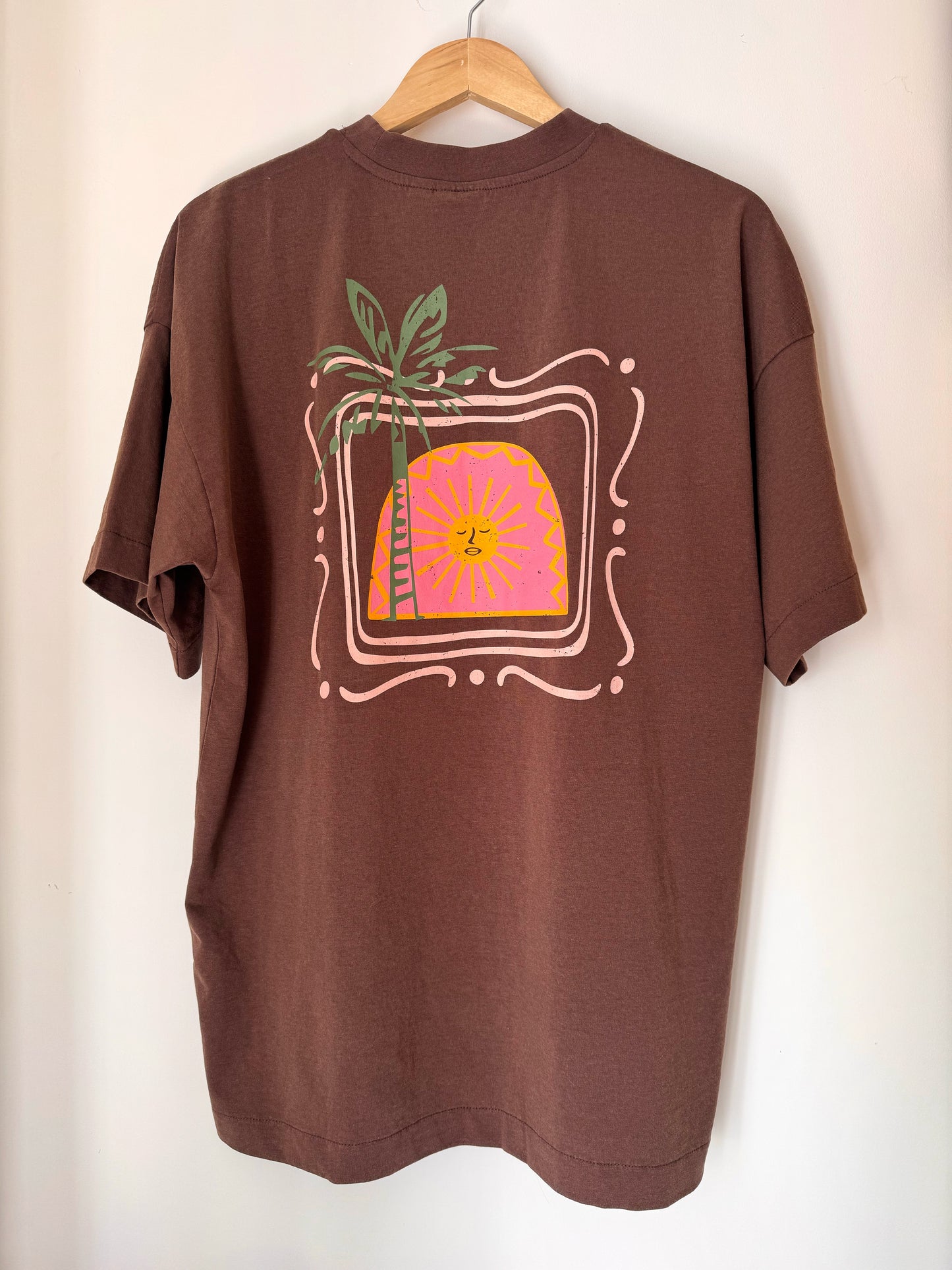 Brown t-shirt with a palm tree and sun design on a hanger against a white wall