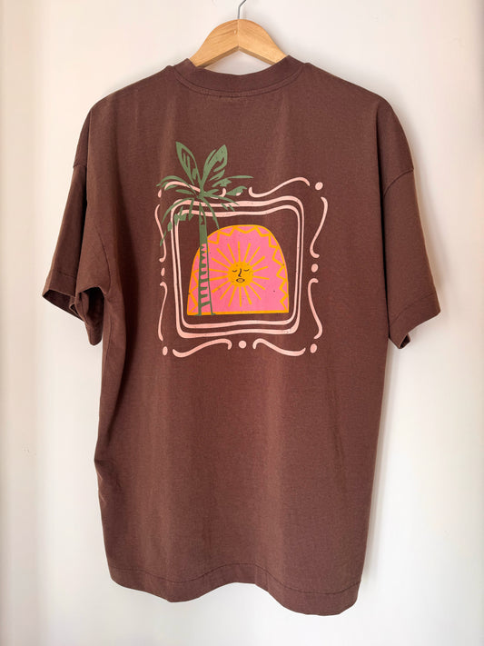 Brown t-shirt with a palm tree and sun design on a hanger against a white wall