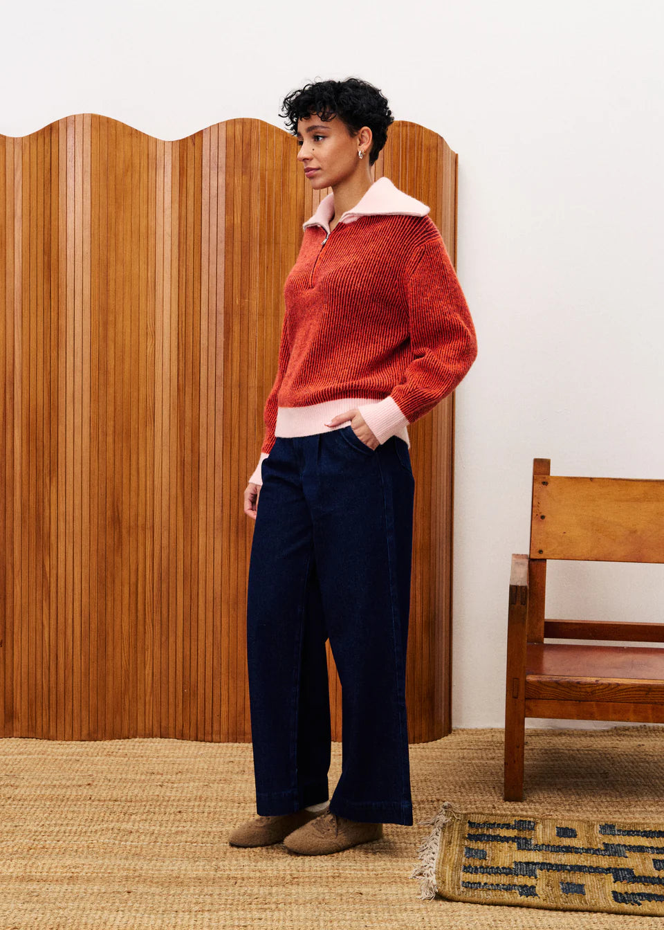 Woman wearing a red sweater and navy pants standing in a room with wooden furniture.